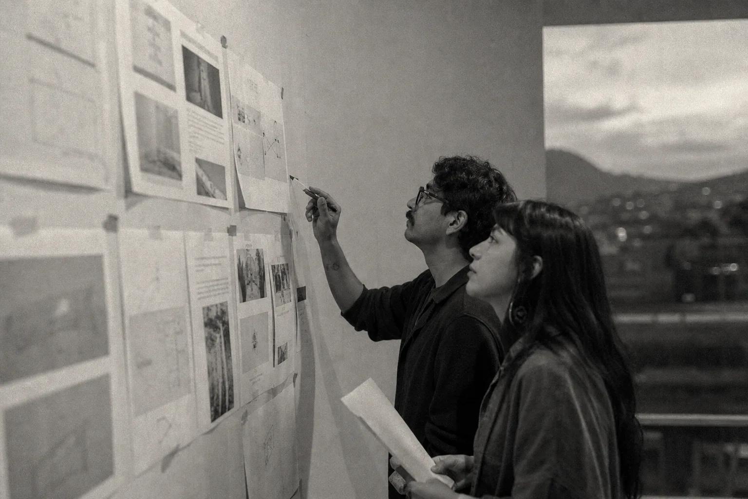 Dos personas revisando trabajo fijado en una pared, con vista a la ciudad, blanco y negro.